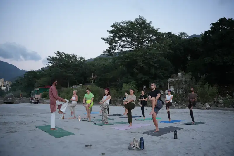 Outdoor yoga class practising balance poses on Ganga beach with Rishikesh hills behind
